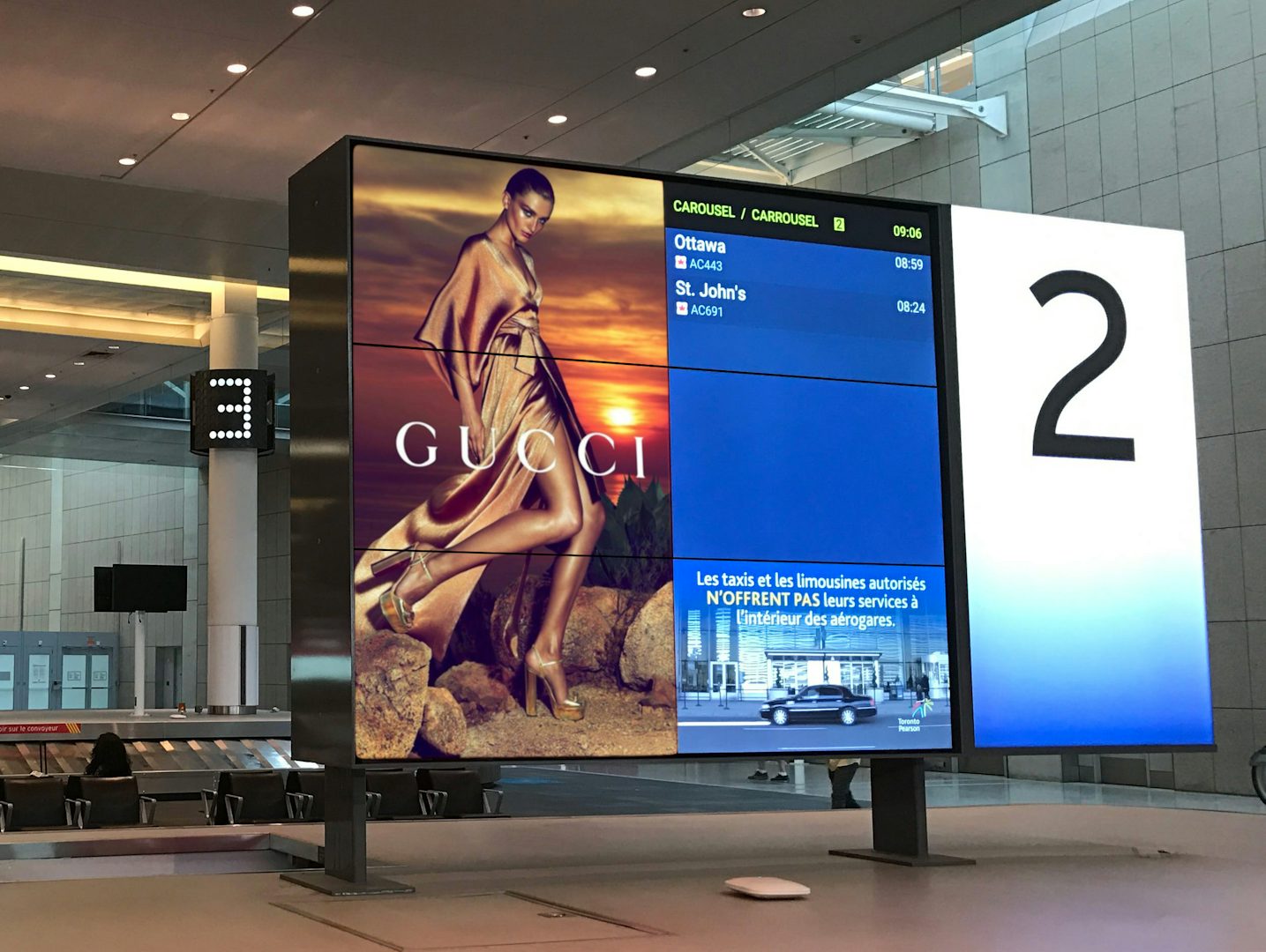 Astral digital board featuring a Gucci ad and flight arrivals at a baggage carousel