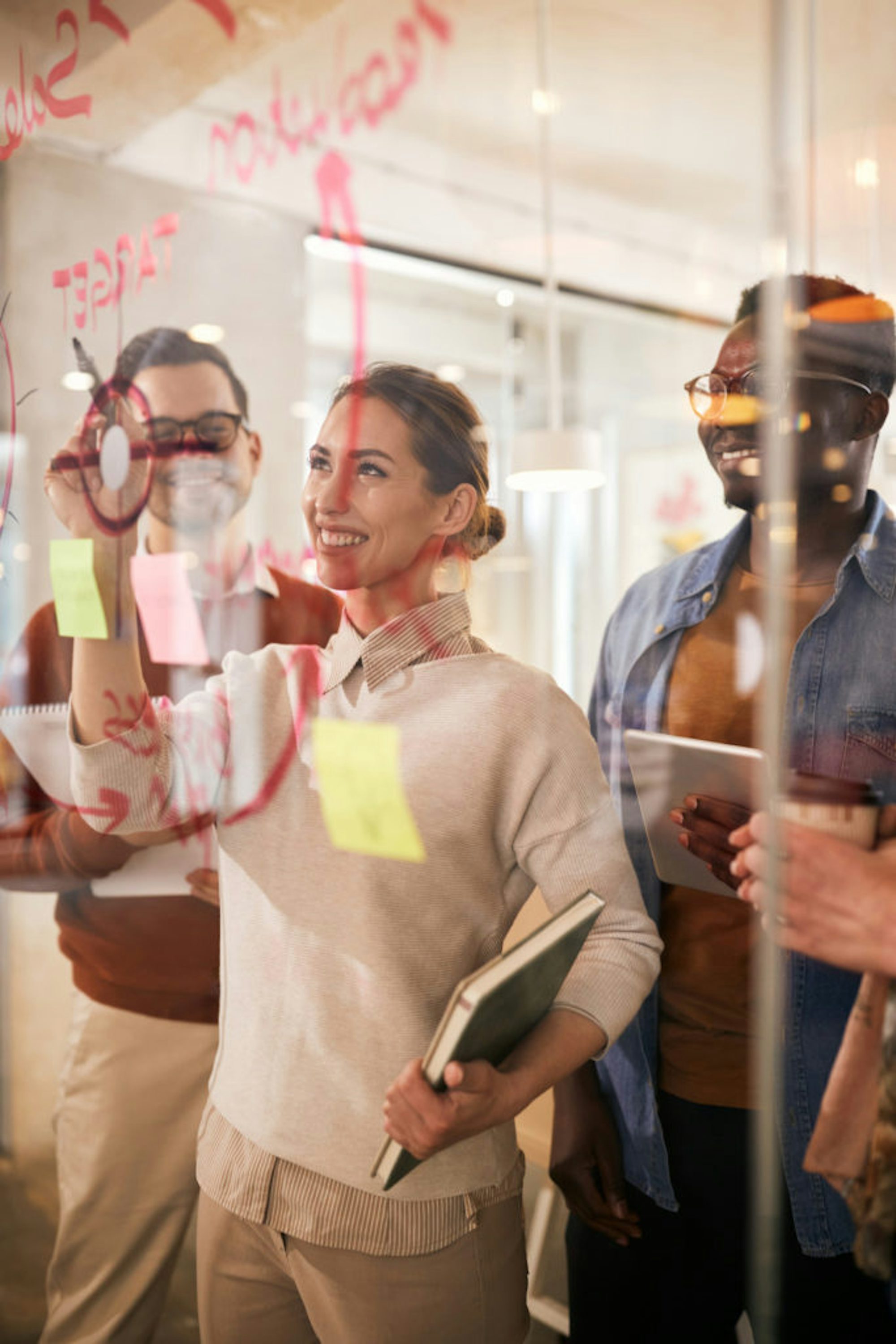 Young happy businesswoman and her colleagues writing plans on glass wall while working in the office. The view is through the glass.