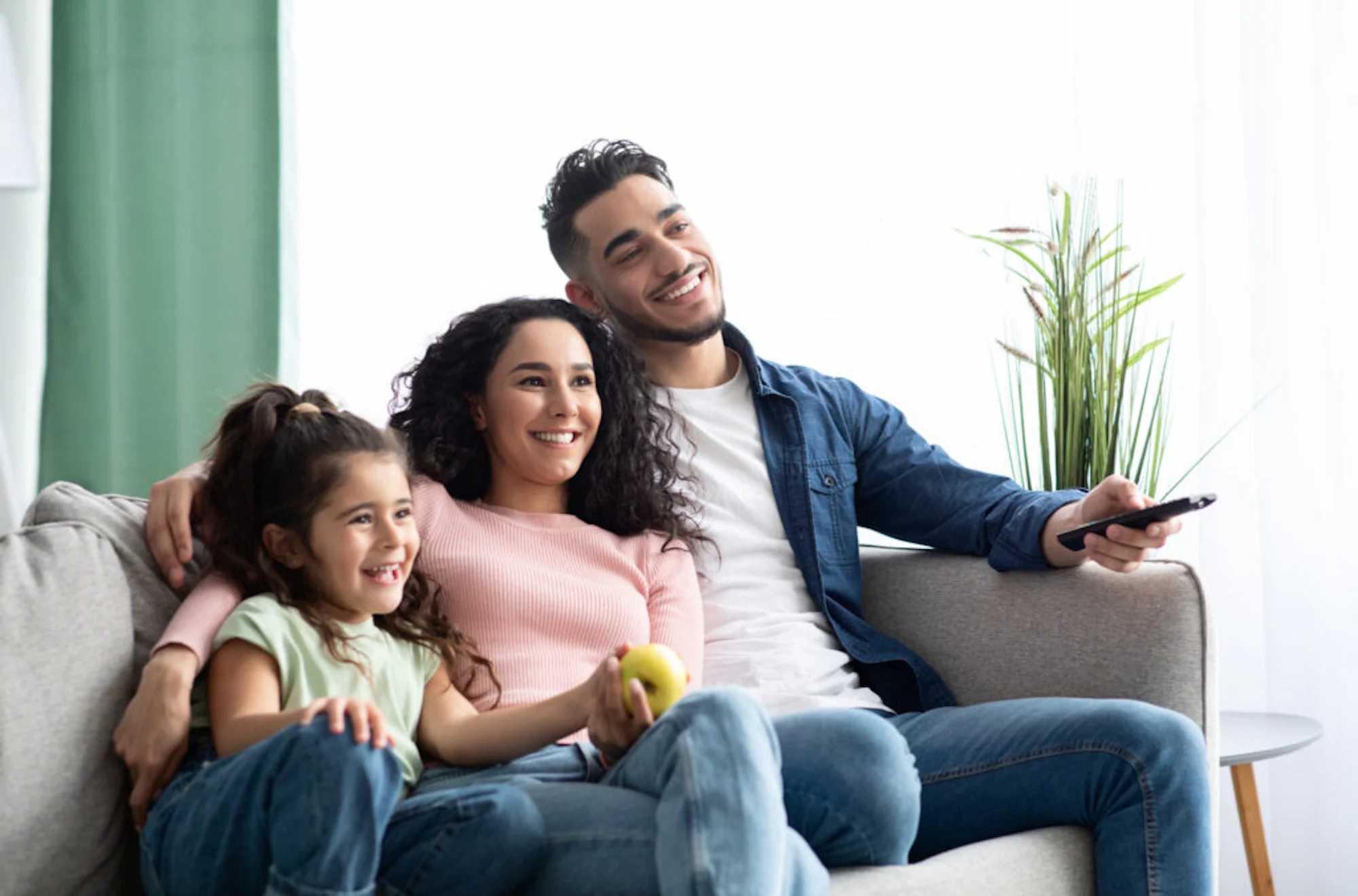 Happy arabic family of three relaxing and watching tv at home, middle eastern father, mother and little daughter resting on couch in living room, enjoying spending weekend together