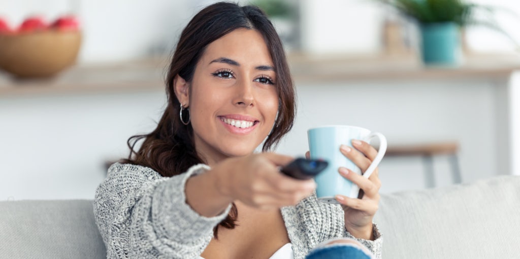 Women holding a coffee cup in one hand and a remote in the other hand. sitting on the couch and looking happy.