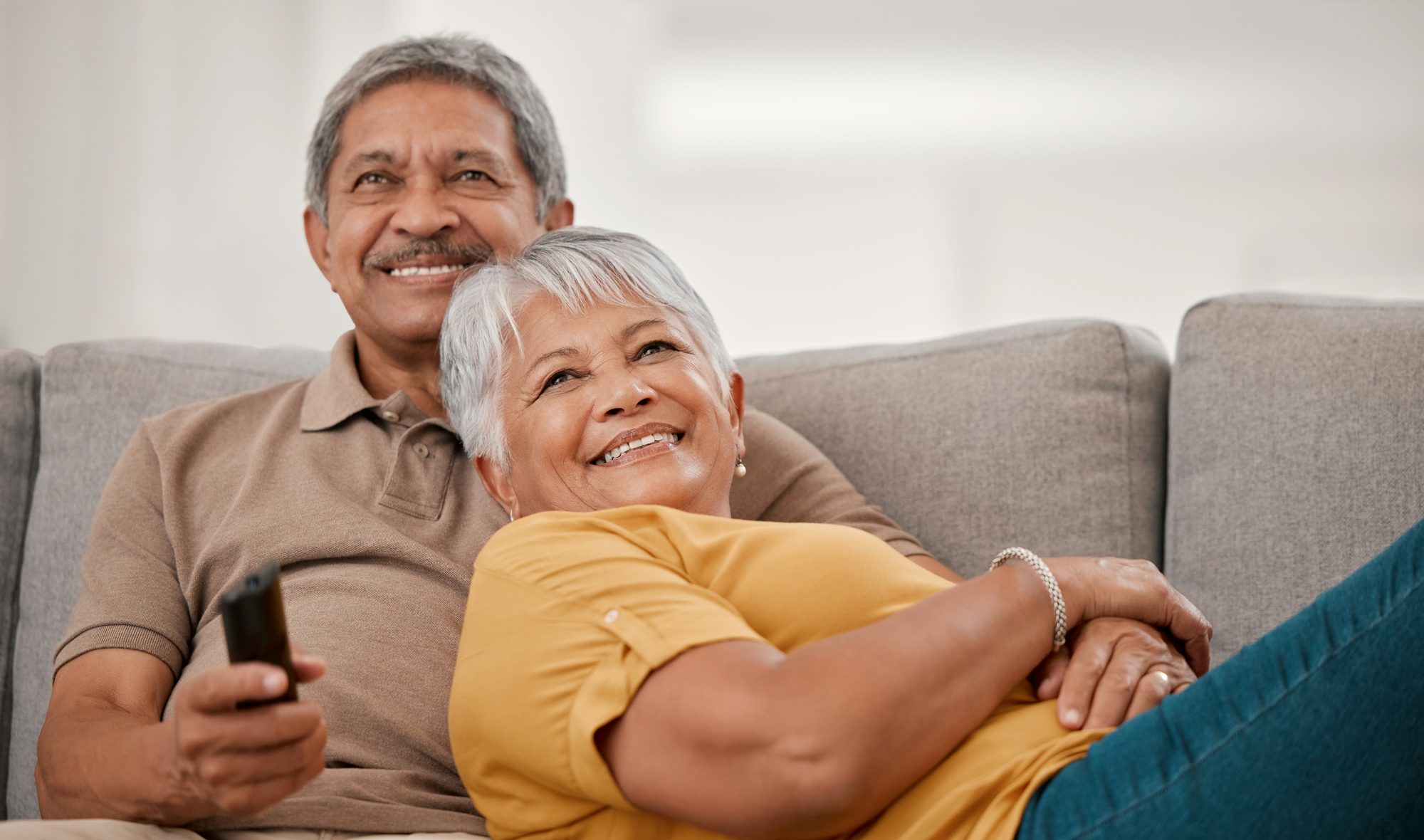 Elderly married man and woman, watching tv on couch bonding, laughing and smiling. The man is holding a remote in one hand.
