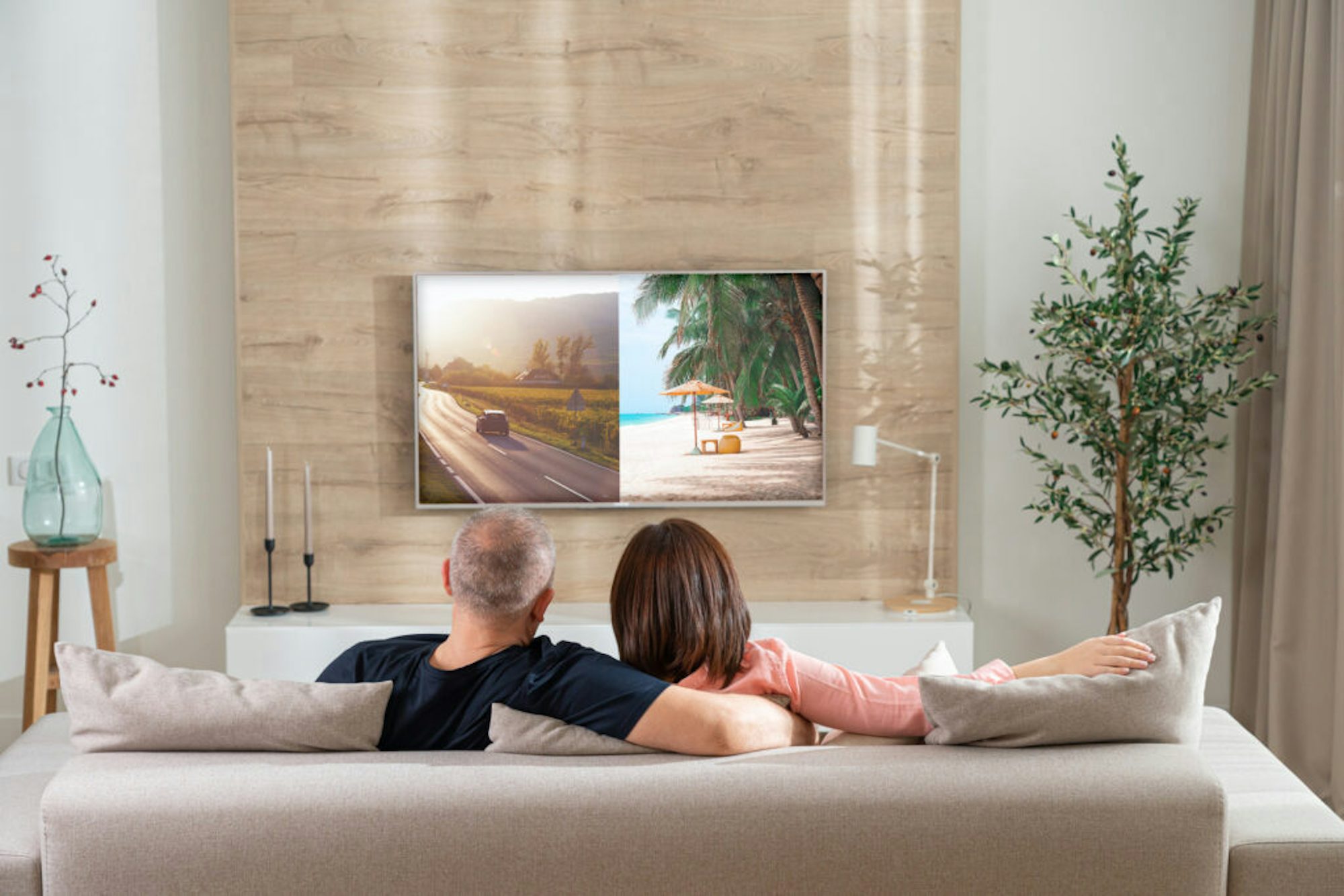 Couple sitting on the couch watching TV. the TV screen is split into 2 One ad is tailored to the men (car) and the other ad is tailored to the women (Beach vacation)