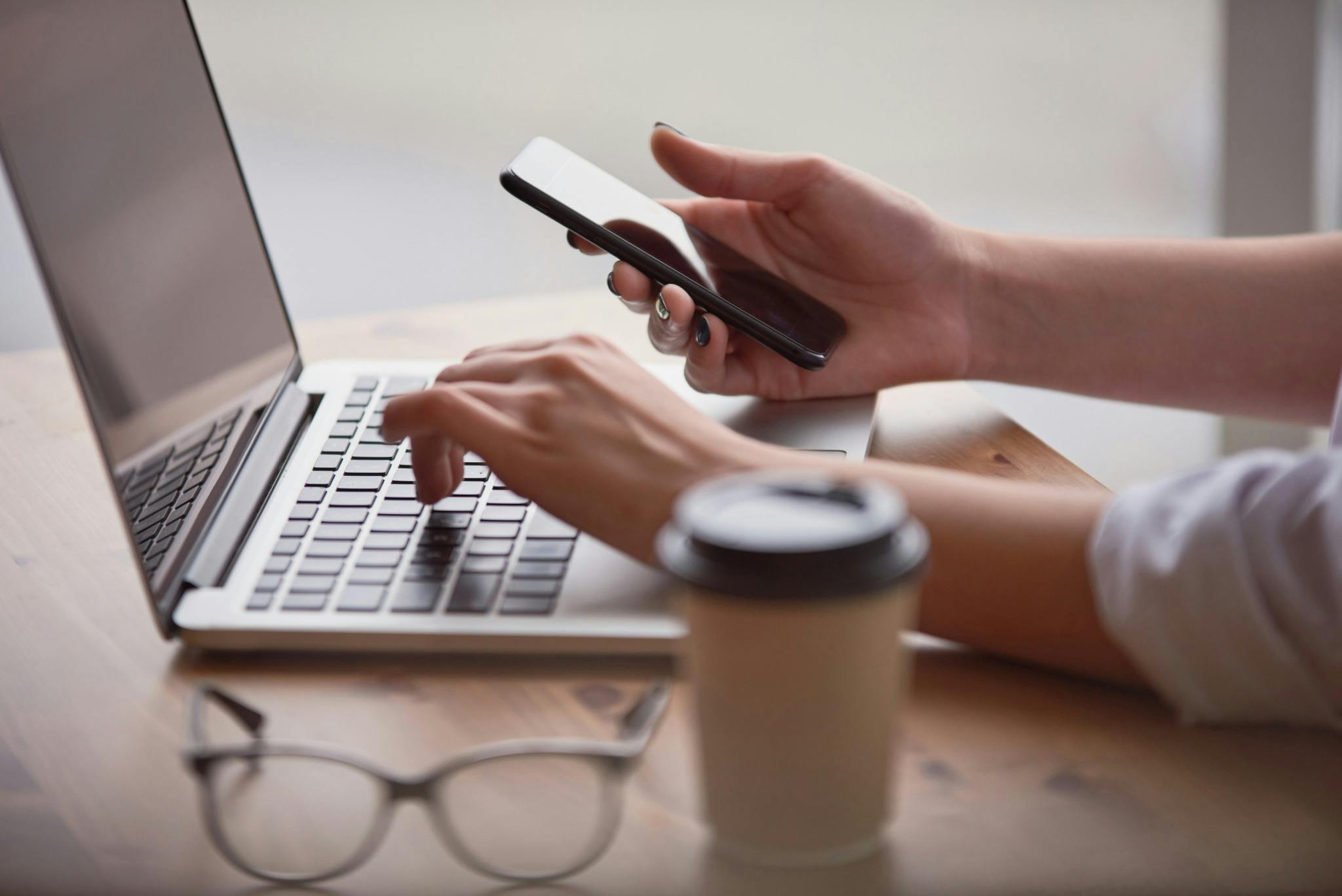 Female hands using smart phone while typing on laptop keyboard at cafe table