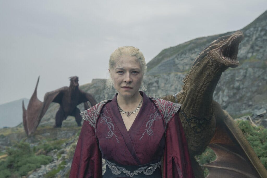 Rhaenyra Targaryen standing in front of two dragons on a mountain.