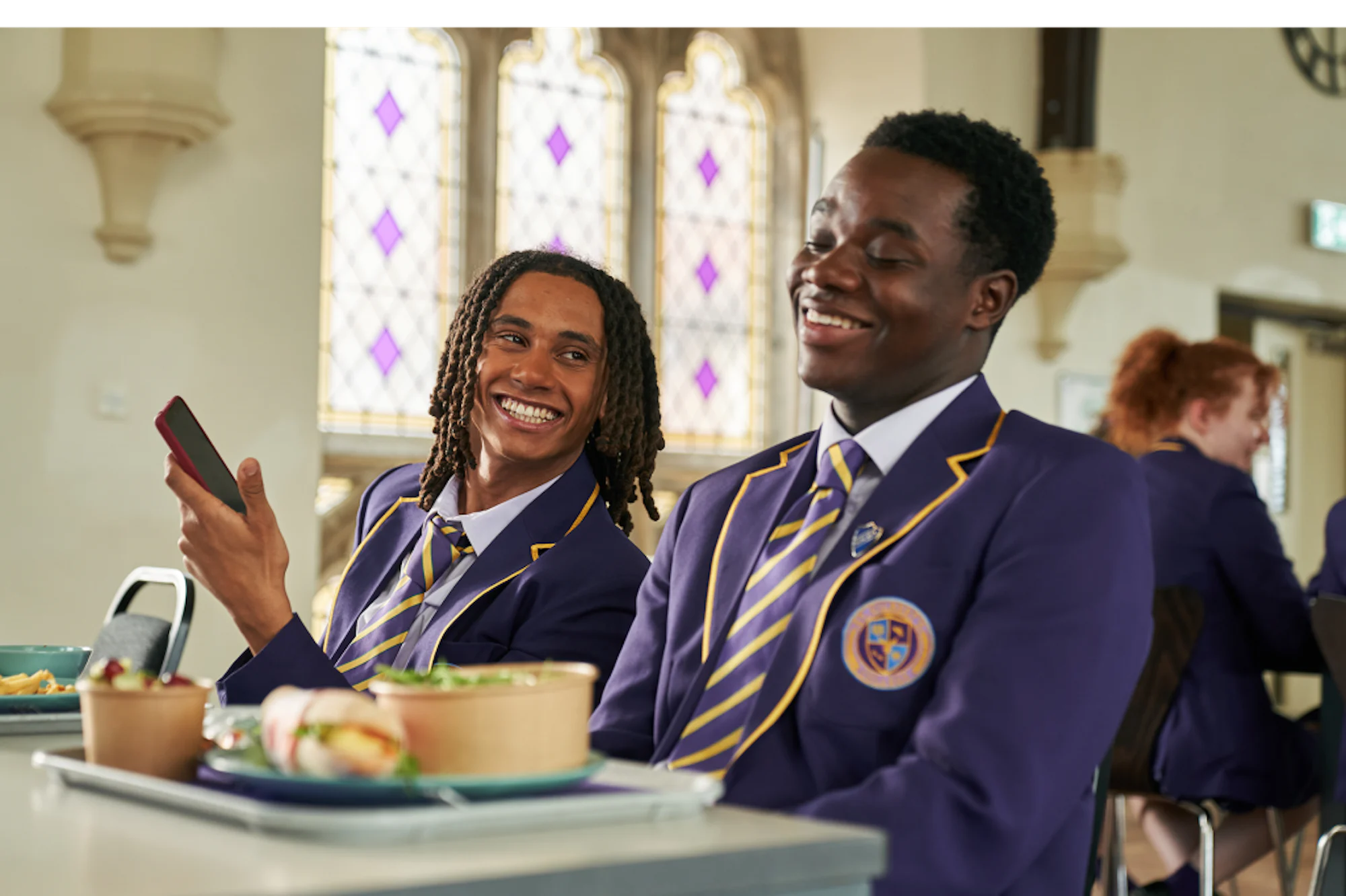 Two students in school uniforms smiling and talking in a cafeteria.