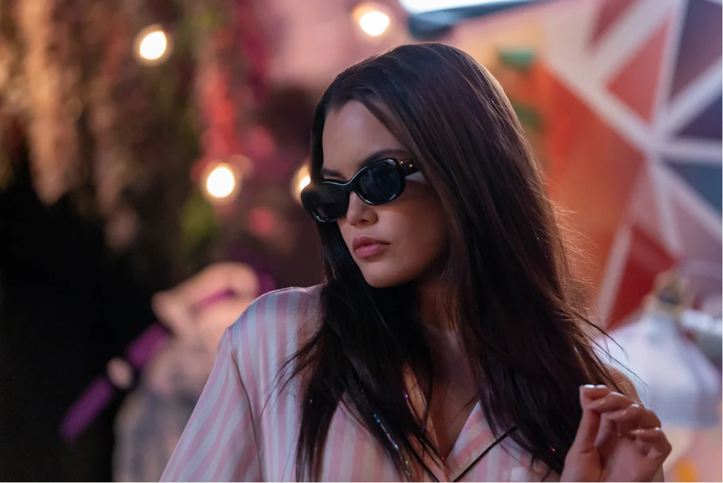 Young woman wearing sunglasses at a colorful outdoor event with lights in the background.