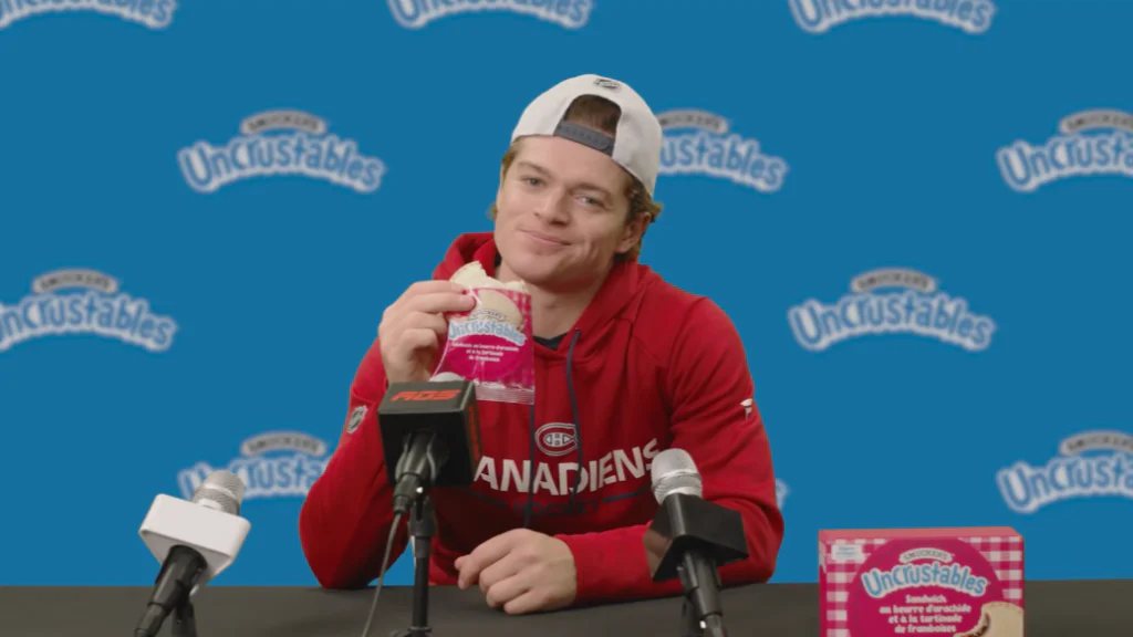 Un joueur de hockey (Cole Caufield) portant un chandail des Canadiens est assis à une table de conférence de presse avec des micros devant lui, tenant un sandwich Uncrustables, avec un fond bleu rempli du logo Uncrustables.